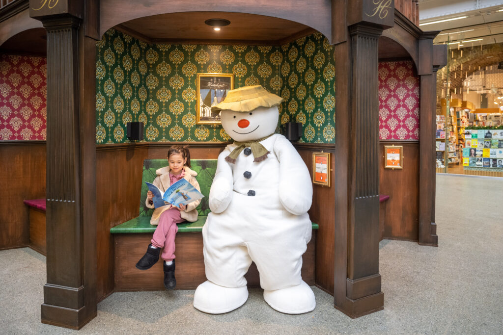Photo of a child reading a book, sitting next to Raymond Briggs' The Snowman, in an audio booth within the St Pancras International Christmas tree